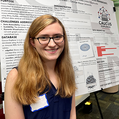 A woman with blondish hair and eyeglasses stands in front of a poster and smiles.