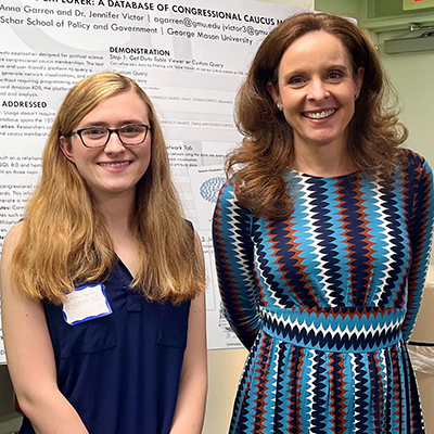 Two women smile at the camera as they stand in front of a poster about political caucuses.