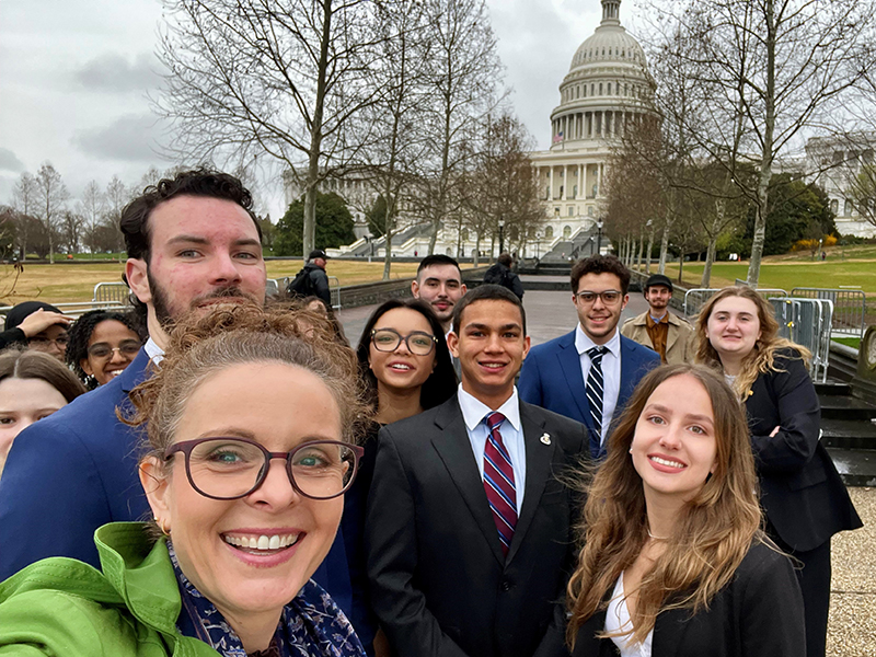 A woman in a green coat takes a selfie with several well dressed young people in front of the U.S. Capitol Building.