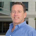 A man in a blue collared shirt stands and smiles