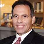 A man in a black suit, white collared shirt, and red tie, stands and smiles