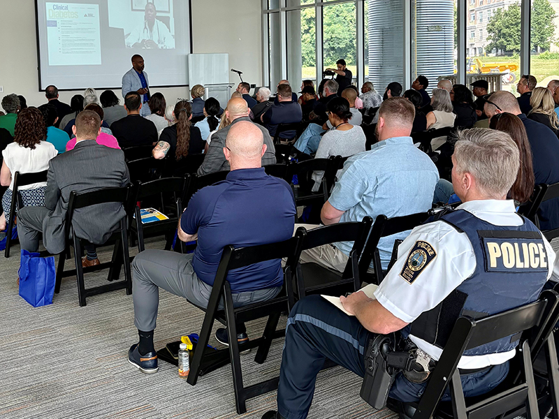 A group of people sit in a conference room setting with one member wearing a jacket reading “police” on the back.