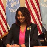 A woman in a black suit and pink shirt sits in front of an American flag and a United Nations flag