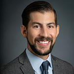 A man in a gray suit, blue tie, and blue collared shirt stands and smiles.