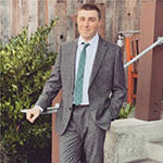 A man in a gray suit, green tie, and white shirt stands outside while smiling