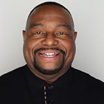 A man in a black shirt stands and smiles