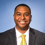 A man in a gray suit, yellow tie, and white collared shirt stands and smiles
