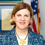 A woman with a jacket and white shirt stands in front of an American flag while smiling