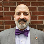 A man in a gray suit, purple bow tie, and blue collared shirt stands smiling in front of a brick wall.