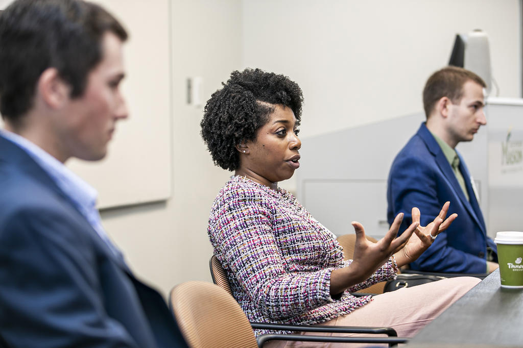 A woman sits speaking in a classroom between two men wearing blue blazers