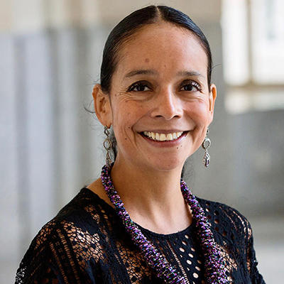A woman wearing a black shirt and purple necklace stands inside and smiles