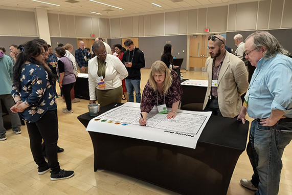 A group of people stand around a table watching a woman write on a large piece of paper
