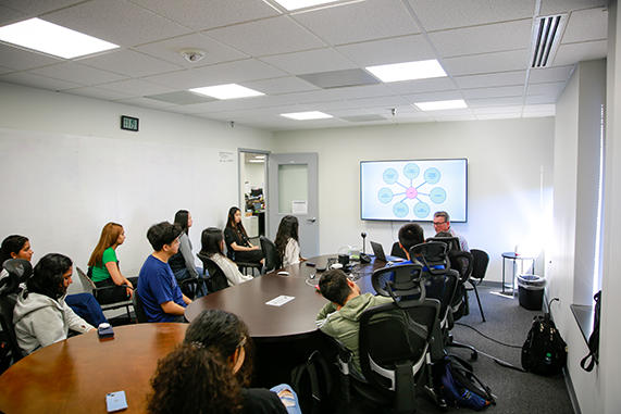 A group of students sit at tables in a classroom while listening to a lecture