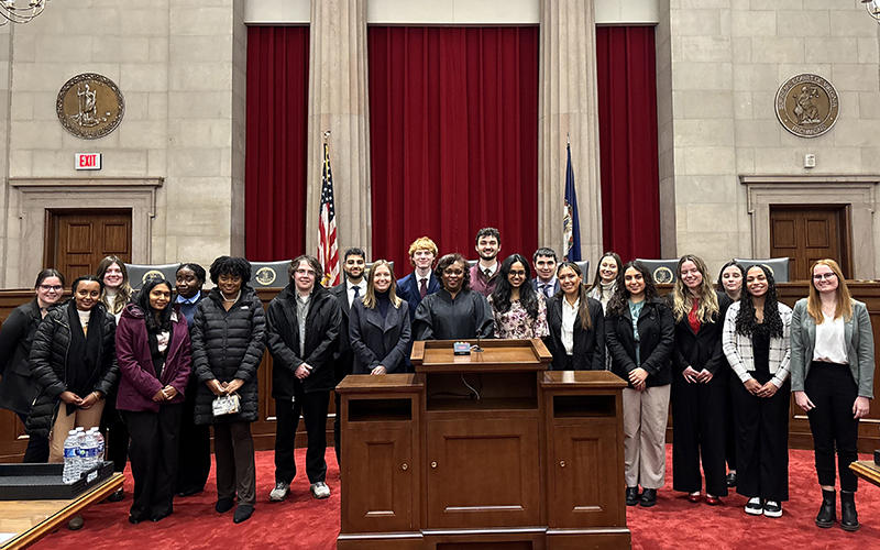 A group of 20 students surround a woman wearing a black robe while standing in front of two columns in a courtroom with a red carpet.