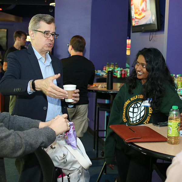 A man in a blue suit stands and speaks to a girl in a green sweater