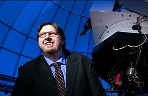 A man in a black suit, blue shirt, and tie stands and smiles