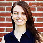 A woman in a blue shirt stands in front of a brick wall and smiles