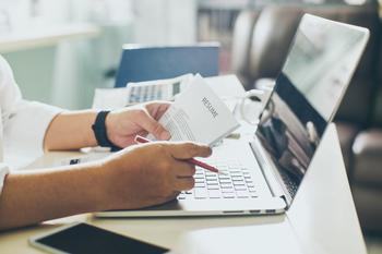 An individual holds a paper with the title, Resume, on it while working on a computer.