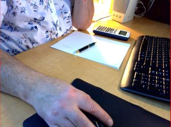 A man's right arm lays on a desk and uses a computer mouse while his left elbow rests on the edge next a a pen on top of a piece of paper.