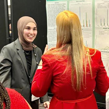 A woman in a green jacket and head scarf talks with a woman in a red outfit.