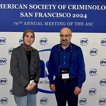 A woman in a green jacket and head scarf stands next to a bald man in a blue shirt in front of a sign for a conference.
