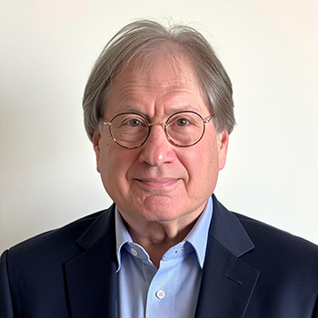A man wearing round-framed glasses and a blue shirt with navy blazer poses for the camera.