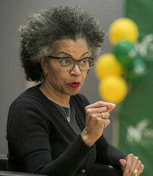 A woman wearing glasses gestures with her right hand to the audience.