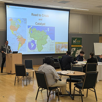 A man in a suit and glasses stands behind a podium behind a screen projecting a world map.