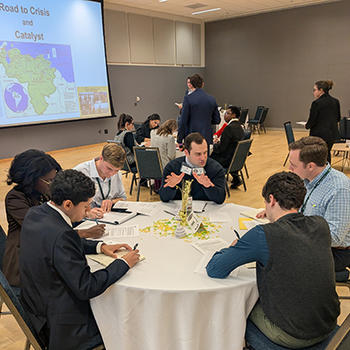 A group of six students sit at a table, surrounded by other tables of students, writing on notepads.