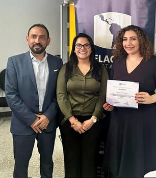 A man in a dark suit jacket stands next to a woman in green and another woman in black holding a certificate.