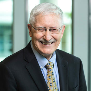 A man in a dark jacket, silver hair, eyeglasses, and a blue shirt smiles at the camera.